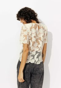 Femme aux cheveux bouclés portant un top en dentelle florale blanche et un jean noir délavé, tournée pour montrer les détails complexes du dos du top.