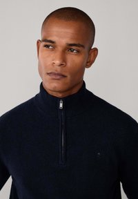 Marineblauer Zip-Pullover aus Strickstoff mit hohem Kragen und dezentem Logodetail auf der Brust. Die Textur erscheint weich und dicht.