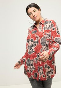 Camicia a bottoni rossa con motivi neri e beige, maniche lunghe e tasca frontale. Abbinata a pantaloni grigi scuri.