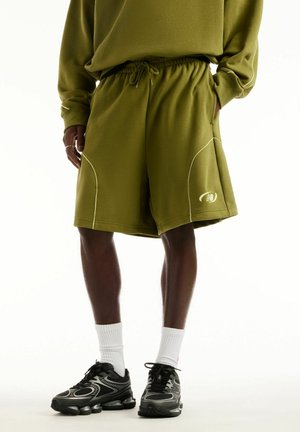 Person trägt olivgrüne Shorts und passendes Sweatshirt, mit weißen Socken und schwarzen Sportschuhen, steht vor weißem Hintergrund.