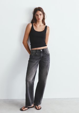 Giovane donna appoggiata a un muro bianco, indossa una canottiera nera, jeans neri sbiaditi e infradito nere, con le mani dietro la schiena.