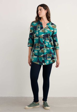 Multicolore tuniek met geometrische patronen in groen, blauw en crème, met een V-hals en driekwart mouwen, gecombineerd met donkere leggings en groene sneakers.