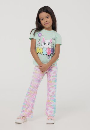 T-shirt in cotone verde chiaro con un gatto fumettistico e testo colorato. Pantaloni a palazzo con motivo tie-dye pastello. Sneakers bianche con dettagli rosa.
