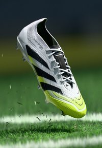 Scarpa da calcio bianca e nera con superficie texture, caratterizzata da tre strisce nere prominenti e un accento giallo brillante sulla suola.
