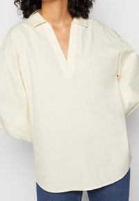 Blouse de couleur crème avec un col en V, des manches courtes et une coupe décontractée. Confectionnée dans un tissu léger à la texture lisse.