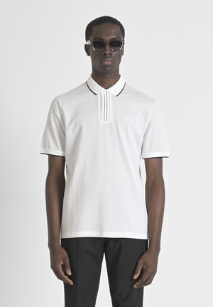 Camisa polo blanca con un tapeta de rayas verticales, detalles en negro en el cuello y las mangas. Usada con pantalones negros.