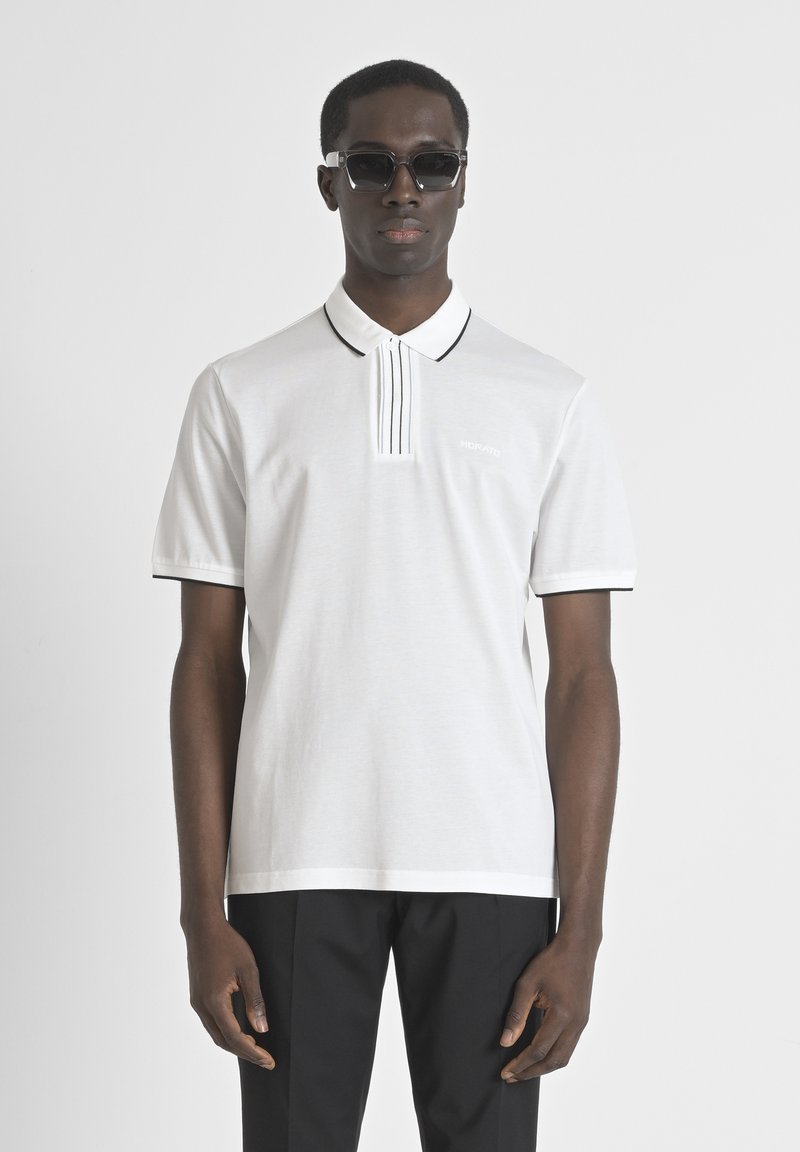 Camisa polo blanca con un tapeta de rayas verticales, detalles en negro en el cuello y las mangas. Usada con pantalones negros.