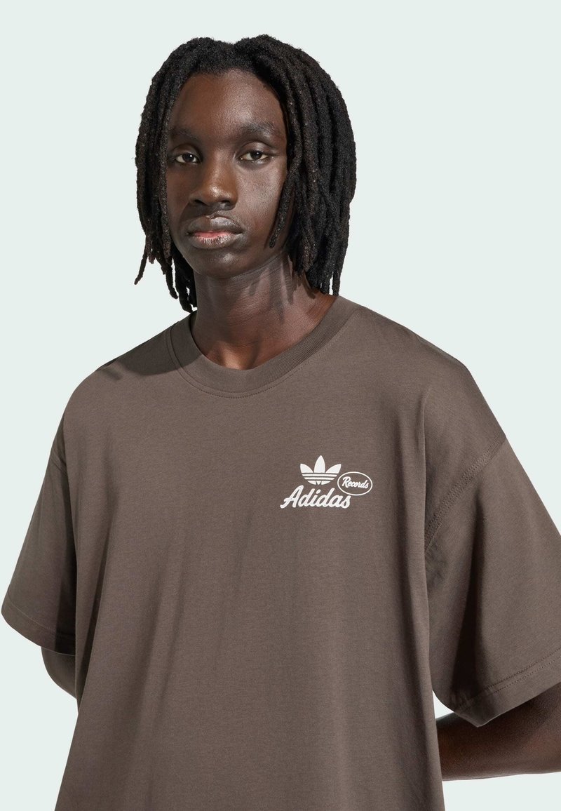T-shirt oversize marrone in morbido cotone, con logo Adidas bianco e scritta sul petto a sinistra. Maniche corte con vestibilità comoda.