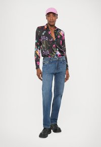 Mulher vestindo uma camisa preta com estampado floral, jeans azuis, sapatos pretos e um boné rosa, de pé contra um fundo branco.