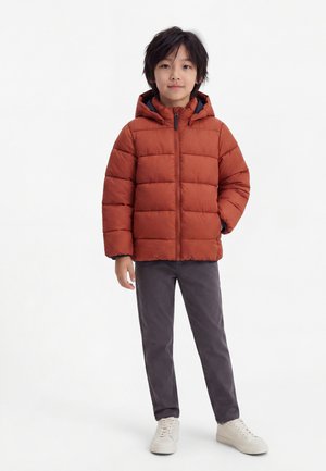 Enfant portant une doudoune couleur rouille avec capuche, pantalon gris et baskets blanches, debout sur un fond blanc uni.