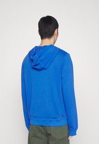 Sudadera con capucha azul, de textura suave y lisa, con cordones ajustables. Presenta puños y dobladillo acanalados, vista desde la parte posterior.