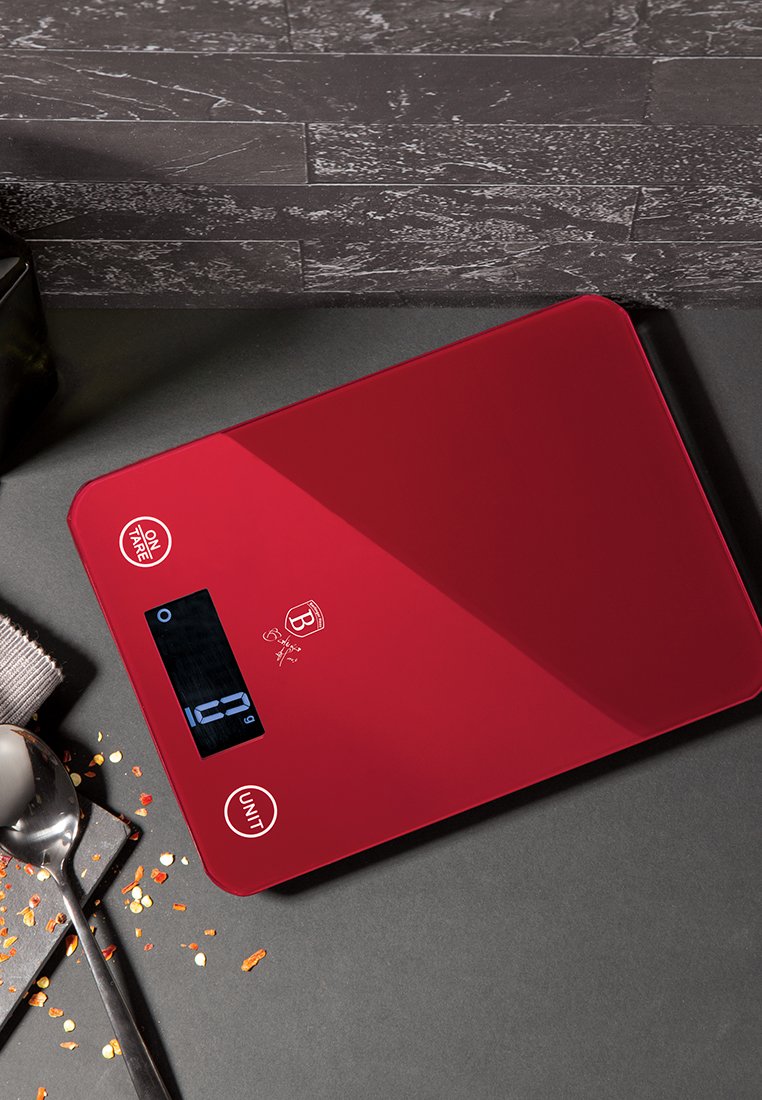 Czerwona cyfrowa waga kuchenna o gładkiej powierzchni, z wyświetlaczem cyfrowym oraz przyciskami oznaczonymi jako "ON TARE" i "UNIT". W pobliżu rozsypane akcenty przypraw.