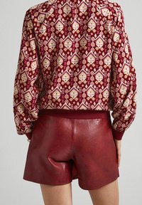 Rode bomberjack met een geometrisch patroon, gecombineerd met wijnrode leren shorts. De texturen tonen een gladde stof en leer.