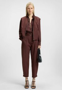Conjunto color burdeos con un chaleco con cremallera y pantalones a juego. Presenta una cintura ceñida, un corte elegante y un pequeño bolso negro. Sandalias de tacón alto con tiras.