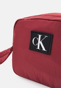 Borsa in nylon rosso con forma rettangolare, caratterizzata da una patch nera che mostra il logo "cK". Texture liscia e chiusura a cerniera visibile.