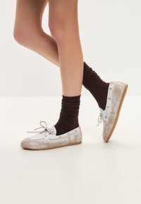 Loafers din velur alb cu sireturi și ocheți maro, asortați cu șosete ribbate maro. Textură netedă și un design cu profil redus.