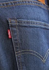 Mörkblå denimjeans har kontrasterande gula sömmar och en liten röd etikett på bakfickan. Dekorativa sömmar längs sidan är synliga.