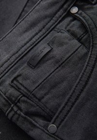 Jeans de mezclilla negra con una textura suave, que presentan una etiqueta de marca visible en el bolsillo y un diseño clásico de cinco bolsillos.