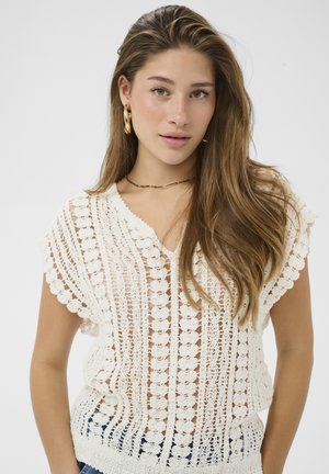 Noor pruuni pikade juustega noor naine, kes kannab valget heegeldatud topi, kuldseid rõngaskõrvarõngaid ja õhukest kaelakeed, poseerides ühevärvilise tausta ees.