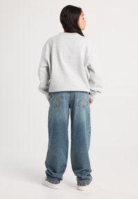 Nuori henkilö seisoo selkä kameraan päin, ja hänellä on päällään vaaleanharmaa collegepaita, löysät siniset farkut ja valkoiset lenkkarit tasaisella taustalla.