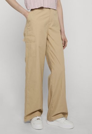 Osoba ubrana w luźne beżowe spodnie typu cargo z dużymi bocznymi kieszeniami, białe trampki oraz krótki różowy top w paski, stojąca na jednolitym tle.