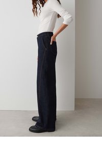 Personne debout de profil, portant une chemise blanche à manches longues, un jean sombre à jambes larges et des chaussures noires, sur un fond intérieur uni.