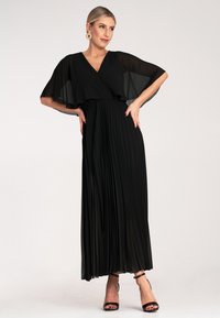 Czarna długa sukienka z plisowanym dołem, przezroczystymi oraz falującymi rękawami i zapinanym na krzyż dekoltem. Stylizowana z czarnymi sandałkami na obcasie i kolczykami w kształcie kół.