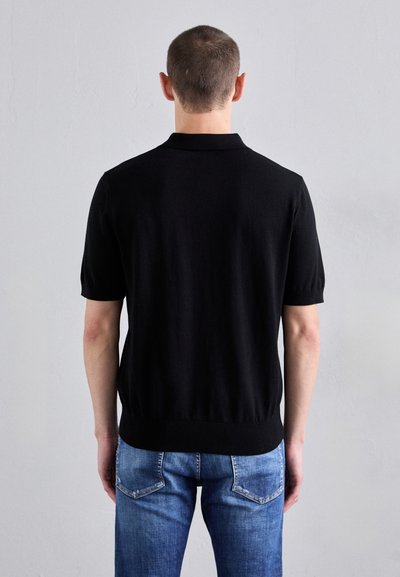 Zwarte polo met korte mouwen van een soepele tricotstof, met een geribde kraag en zoom, gedragen met lichtblauwe denim jeans.