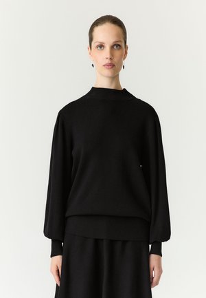Femme aux cheveux plaqués en arrière, portant un pull noir à manches longues et une jupe, debout devant un fond blanc uni.