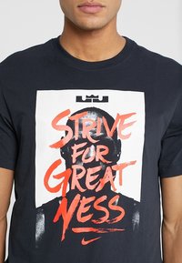 Camiseta de algodón negra con un gran gráfico blanco que presenta una silueta y un texto rojo en negrita que dice "ESFUÉRZATE POR LA GRANDIOSIDAD". Logotipo de Nike mostrado.