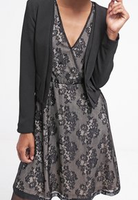 Blazer noir porté sur une robe en dentelle florale grise, avec un décolleté en V profond, des manches courtes et une superposition en dentelle transparente à ourlet festonné.