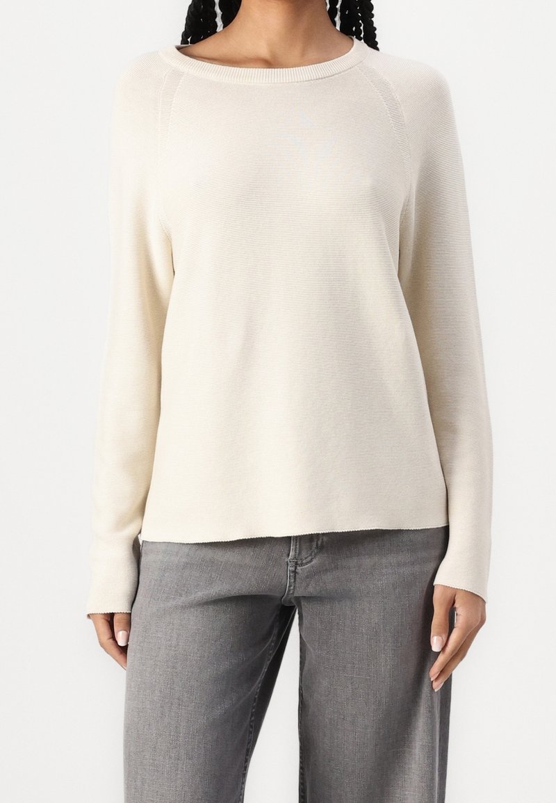 Light beige long-sleeve sweater with a round neckline, ribbed texture, and slightly loose fit. Paired with gray denim pants.