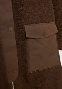 Hnědý kabát s texturovaným chlupatým materiálem a hladkým látkovým panelem, s kapsou na klopu se zacvakávacím knoflíkem. Obsahuje také detail s kovovým zipem.