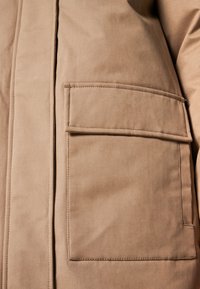 Brun, tekstureret stofjakke med en fremtrædende lomme, lodret søm og kontrasterende stikning. Glat overflade, praktisk design.
