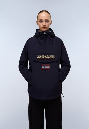 Giovane donna con giacca Napapijri blu navy con collo alto con zip e patch della bandiera norvegese, mani nelle tasche, sfondo neutro.