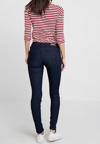 Kvinna klädd i mörkblå tajta jeans, röd och vit randig topp med trekvartslånga ärmar och vita sneakers, står vänd bort.