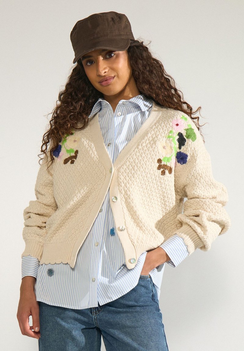 Femme aux cheveux bouclés portant une casquette marron, un cardigan en maille crème avec des broderies florales, une chemise rayée bleue et un jean bleu, posant la main dans la poche.