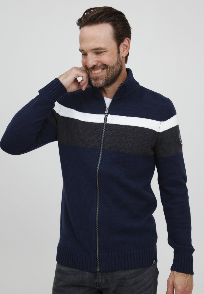 Sudadera de cremallera en navy con un cuello acanalado, que presenta rayas blancas y grises en el pecho. Textura de punto, mangas largas y diseño ajustado.