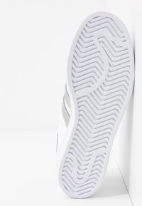 Bílá podrážka tenisky se zvlněným a zakřiveným vzorem, teniska nakloněná proti bílé zdi a rohu podlahy.