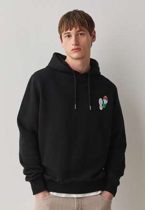 Junger Mann trägt einen schwarzen Hoodie mit kleinen bunten Fußabdruck-Designs auf der Brust, steht vor einer hellen Wand mit den Händen in den Taschen.