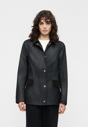 Vrouw met krullend bruin haar, gekleed in een zwarte jas met knopen en klepzakken over een wit shirt, staand tegen een eenvoudige lichte achtergrond.