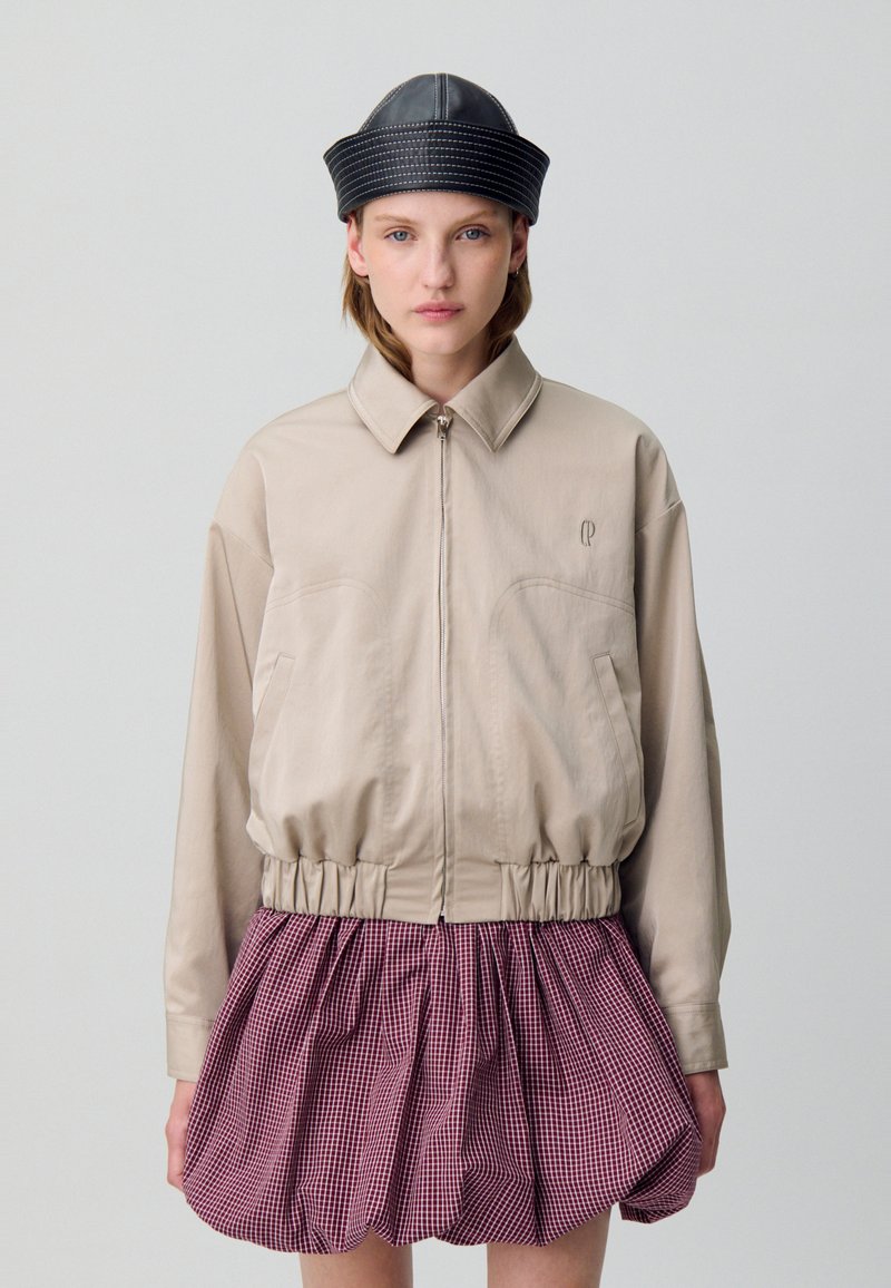 Giacca beige con zip e vita cintrata, abbinata a una gonna a scacchi rossa e bianca. Un cappello di pelle nera completa l'outfit.