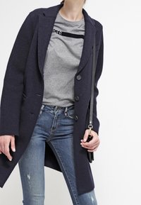 Laivastonsininen villasekoitepäällystakki, jossa on pitkä siluetti, suuret kaulukset ja napitus, yhdistetty harmaaseen t-paitaan ja kulutettuihin sinisiin farkkuihin.