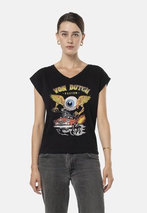 Femme portant un t-shirt noir Von Dutch avec un graphisme d'un globe oculaire ailé et une voiture rouge en flammes, associé à un jean gris et des boucles d'oreilles créoles dorées.