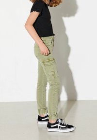 Pantalones cargo verde claro con múltiples bolsillos, corte ajustado y dobladillo fruncido. Combinados con zapatillas negras y una camiseta negra de canalé.