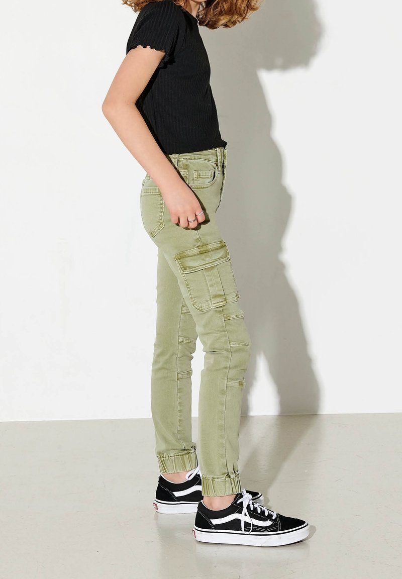 Pantalones cargo verde claro con múltiples bolsillos, corte ajustado y dobladillo fruncido. Combinados con zapatillas negras y una camiseta negra de canalé.