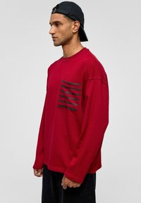 Rode sweatshirt met een grote zwarte gestreepte zak, lange mouwen en een losse pasvorm. De stof is zacht met een gestructureerde oppervlakte.