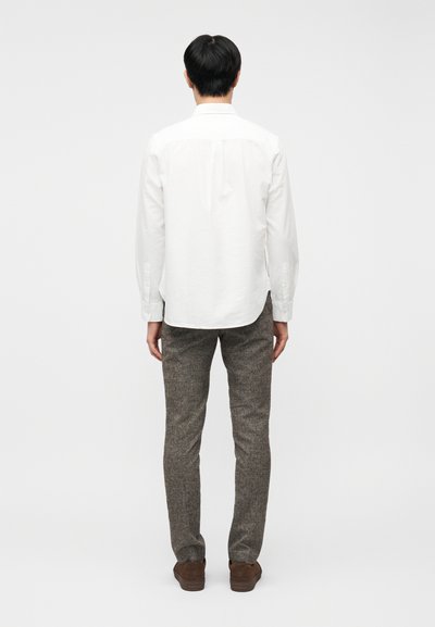 Άνδρας με άσπρο πουκάμισο μακριά μανίκια, γκρι παντελόνι τουίντ και καφέ παπούτσια, που στέκεται γυρισμένος ανάποδα μπροστά σε απλό φωτεινό φόντο.