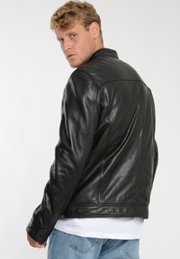 Sort læderjakke med en glat tekstur, der har en spids krave og synlige sømme. Båret over en hvid skjorte, modellen vender ryggen til.