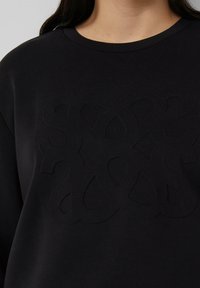 Czarna bluza z teksturowanym, wypukłym wzorem przedstawiającym splecione kształty. Klasyczny fason z okrągłym dekoltem, miękki materiał, minimalistyczny styl.